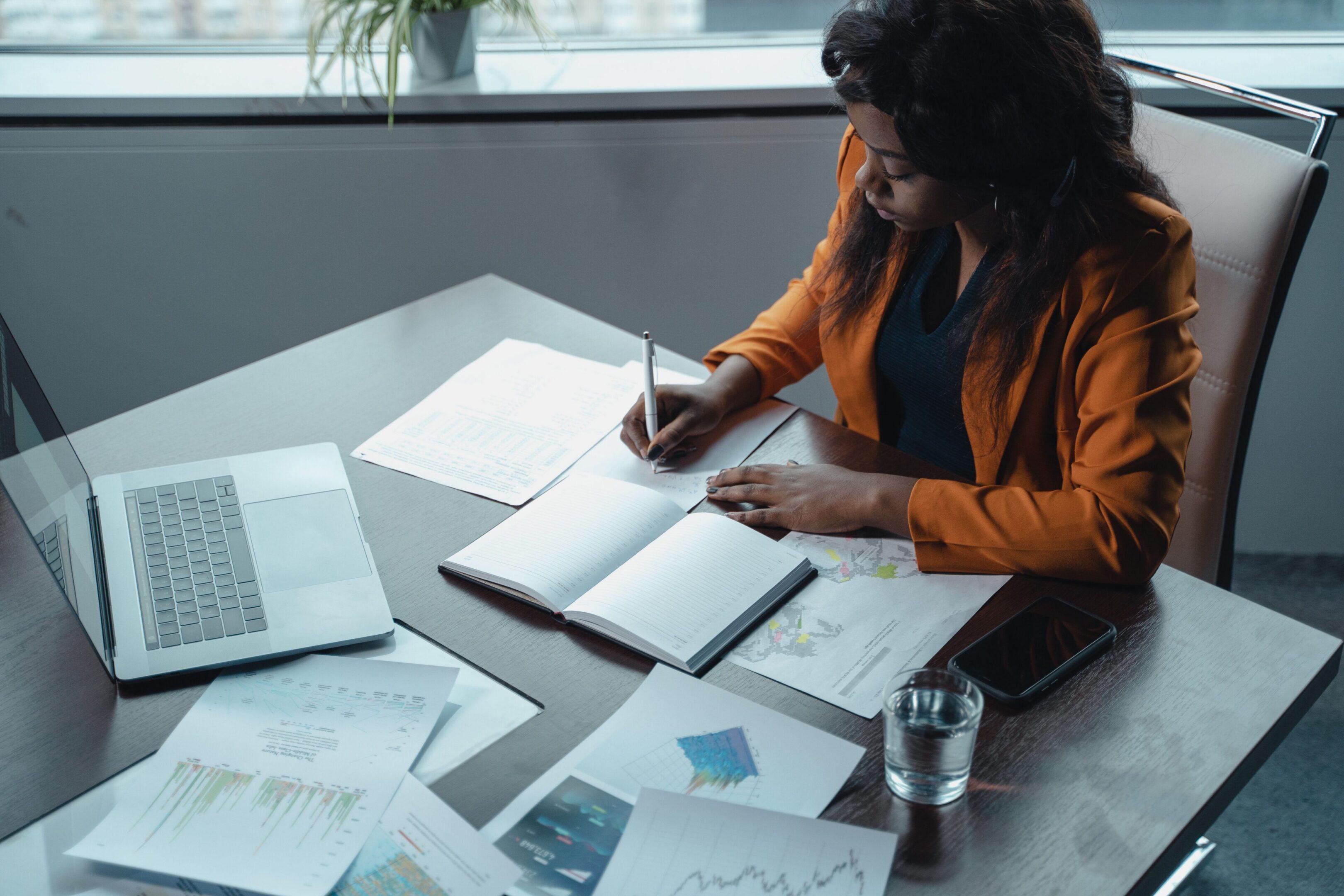 Professional businesswoman reviewing documents and taking notes at a desk in a modern office with a laptop, reports, and business charts.